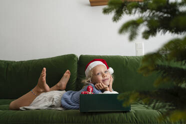 Lächelndes Mädchen auf dem Sofa liegend mit einer Schachtel Weihnachtsschmuck - SVKF01625