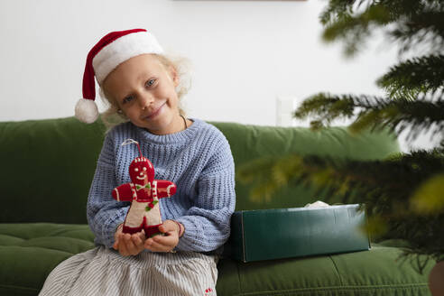 Smiling girl holding Christmas decoration on sofa at home - SVKF01626