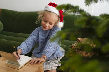 Cheerful girl writing letter to Santa and sitting on sofa at home - SVKF01630