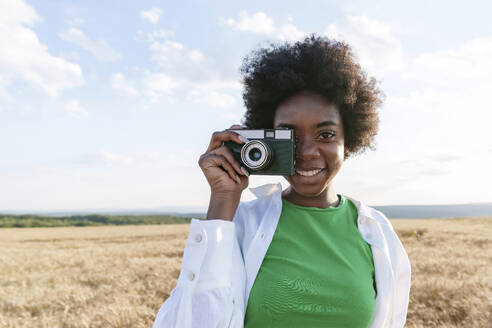 Happy woman with curly hair photographing through camera in field - AAZF01086