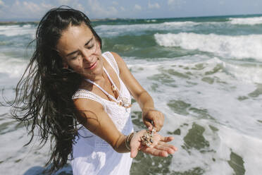 Frau mit langen Haaren hält Muscheln in der Hand in der Nähe von Meer am Strand - SIF00950