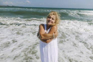 Smiling senior woman hugging self standing near sea at beach - SIF00958