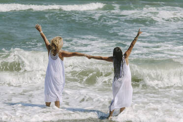 Unbeschwerte Freunde mit erhobenen Händen im Meer am Strand stehend - SIF00967