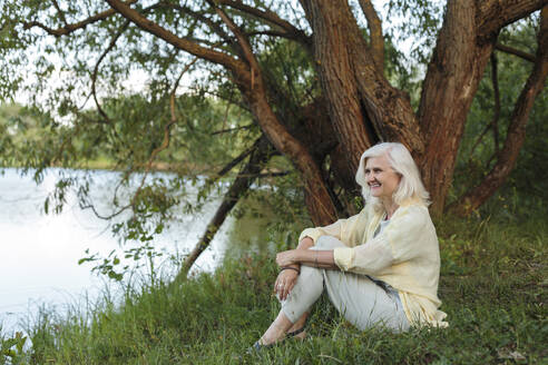Happy mature woman sitting at lakeshore - LLUF01068