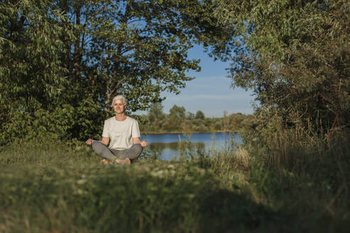 Mature woman practicing lotus position and meditating at lakeshore - LLUF01079