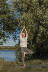 Mature woman practicing tree pose yoga on mat at lakeshore - LLUF01080