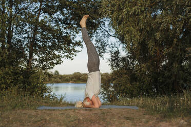 Mature woman practicing headstand yoga on mat at lakeshore - LLUF01082