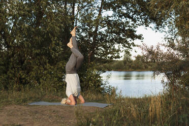 Mature woman practicing headstand yoga on mat at lakeshore - LLUF01083
