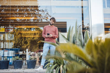 Smiling man with smart phone standing in front of glass wall - JOSEF21055