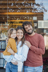 Smiling man with arm around woman and daughter in front of glass wall - JOSEF21067