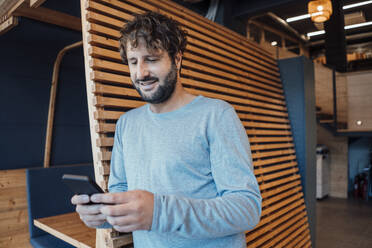 Businessman using mobile phone leaning on wooden wall at office - JOSEF21082