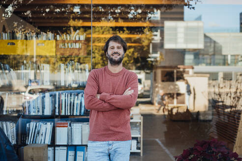 Smiling man standing with arms crossed in front of glass wall - JOSEF21138