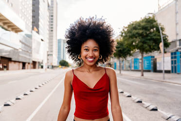 Eine junge, glückliche afrikanische Frau mit Afro-Lockenfrisur genießt einen Spaziergang in der Stadt - eine fröhliche schwarze Studentin unterwegs - DMDF05967