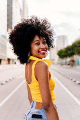 Junge, glückliche afrikanische Frau mit Afro-Lockenfrisur genießt einen Spaziergang durch die Stadt - fröhliche schwarze Studentin unterwegs - DMDF05975