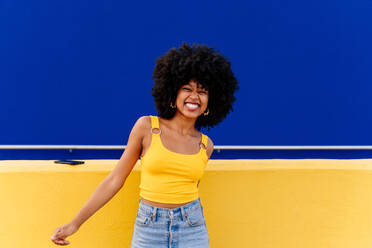 Junge, glückliche afrikanische Frau mit Afro-Lockenfrisur genießt einen Spaziergang in der Stadt - Porträt einer fröhlichen schwarzen Studentin vor einem bunten Wandhintergrund - DMDF05992