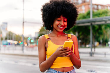 Junge, glückliche Afro-Frau mit Lockenfrisur genießt einen Spaziergang durch die Stadt - fröhliches schwarzes Studentenmädchen auf der Straße - DMDF06035