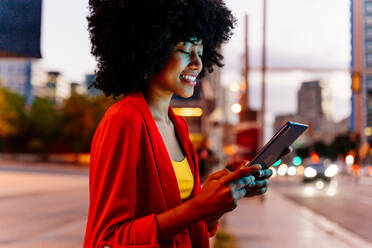 Junge, glückliche Afro-Frau mit Lockenfrisur genießt abendlichen Spaziergang durch die Stadt - ein fröhliches Studentenmädchen - DMDF06048