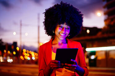 Junge, glückliche afrikanische Frau mit Afro-Lockenfrisur genießt einen abendlichen Spaziergang in der Stadt - ein fröhliches schwarzes Studentenmädchen - DMDF06049