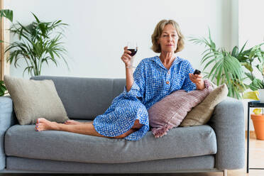 Beautiful mature senior woman at home, domestic life and leisure moments - 50-60 years old pretty female adult relaxing on the sofa and drinking wine while watching tv - DMDF06318