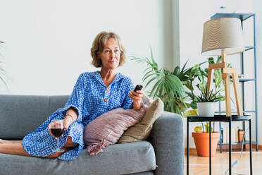 Eine entspannte ältere Frau genießt ihr häusliches Leben und Freizeitmomente auf dem Sofa mit einem Glas Wein und Fernsehen - DMDF06319