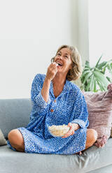 Eine entspannte ältere Frau genießt ihr Zuhause und ihre Freizeit, indem sie auf dem Sofa sitzt, einen Film schaut und Popcorn isst - DMDF06320