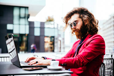 Junger Hipster-Mann arbeitet an einem Laptop in einem Café im Freien - DMDF06526