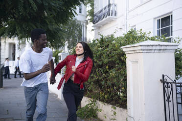 Fröhliches junges Paar hält sich an den Händen und läuft in der Nähe eines Gebäudes - AMWF01728