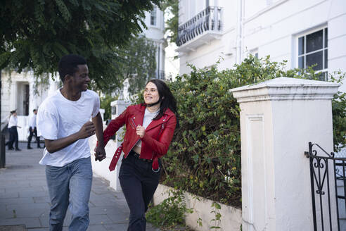 Cheerful young couple holding hands and running near building - AMWF01728