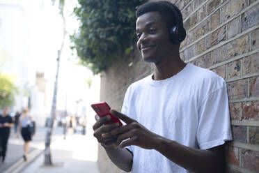 Lächelnder junger Mann, der drahtlose Kopfhörer trägt und mit seinem Handy an der Wand steht - AMWF01743
