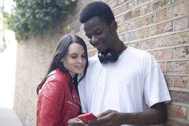 Smiling couple using smart phone near wall - AMWF01744