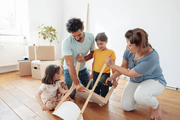 Woman using drill machine on wooden chair with family in front of wall - JOSEF21219