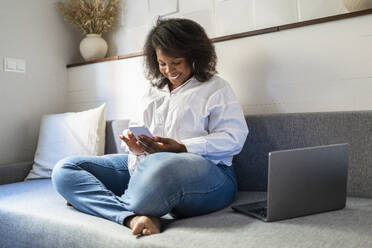 Lächelnder Freiberufler sitzt mit Laptop und Smartphone auf dem Sofa zu Hause - DLTSF03649