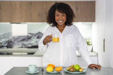 Smiling woman with orange juice having breakfast at home - DLTSF03651