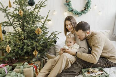 Junge Eltern schenken ihrer kleinen Tochter auf dem Bett zu Hause Weihnachtsschmuck - VBUF00414