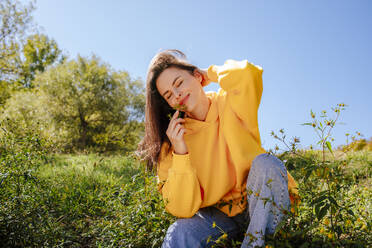 Happy woman sitting near plants and smelling flower on sunny day - MDOF01515