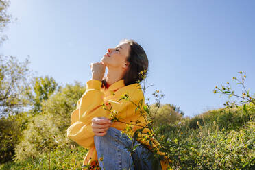 Smiling woman with hand on chin sitting in nature under sky - MDOF01518