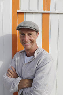 Happy man with arms crossed standing in front of wooden house - GISF00974