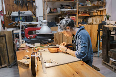 Handwerkerin bei der Arbeit an einer Geige am Schreibtisch in einer Werkstatt - MMPF00971