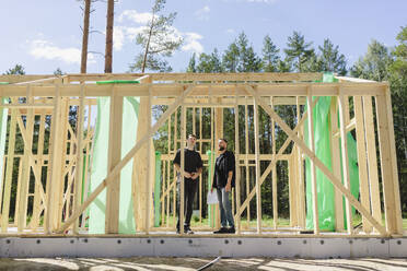 Ingenieure stehen mit Blaupause in der Nähe von Holzrahmen an einem sonnigen Tag - SEAF02045