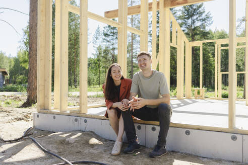 Lächelndes Paar sitzt in der Nähe von Holzkonstruktionen an einem sonnigen Tag - SEAF02059