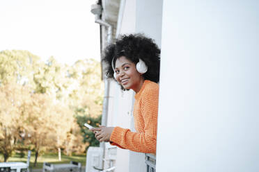 Lächelnde Frau mit drahtlosen Kopfhörern und Smartphone auf dem Balkon - MDOF01556
