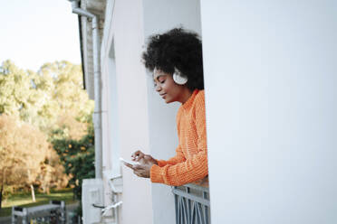 Smiling woman wearing wireless headphones using smart phone in balcony - MDOF01557