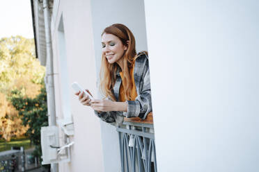 Smiling redhead woman using smart phone in balcony - MDOF01564