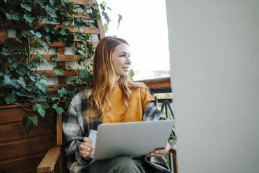 Smiling redhead woman sitting with credit card and using laptop in balcony - MDOF01569