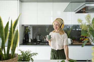 Lächelnde Frau mit drahtlosen Kopfhörern, die eine Kaffeetasse in der Küche zu Hause hält - SVKF01645