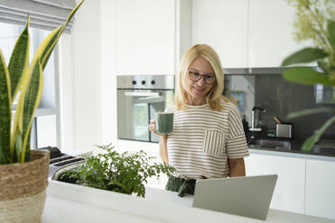 Smiling freelancer with coffee cup working on laptop at home - SVKF01650