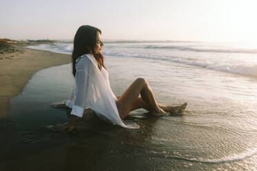 Lächelnde reife Frau sitzt in der Nähe von Meer am Strand - SIF01022