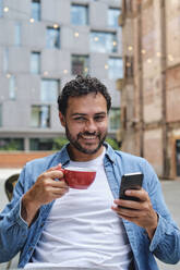 Lächelnder Mann sitzt mit Kaffeetasse und Smartphone vor einem Gebäude - ASGF04642
