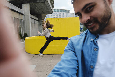 Smiling man taking selfie with woman jumping in background - ASGF04648