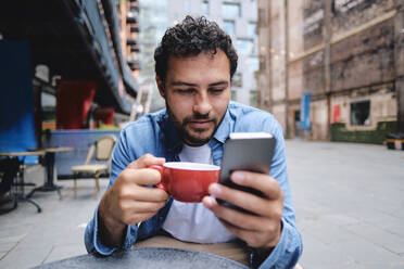Mann hält Kaffeetasse und benutzt Smartphone in einem Straßencafé - ASGF04663